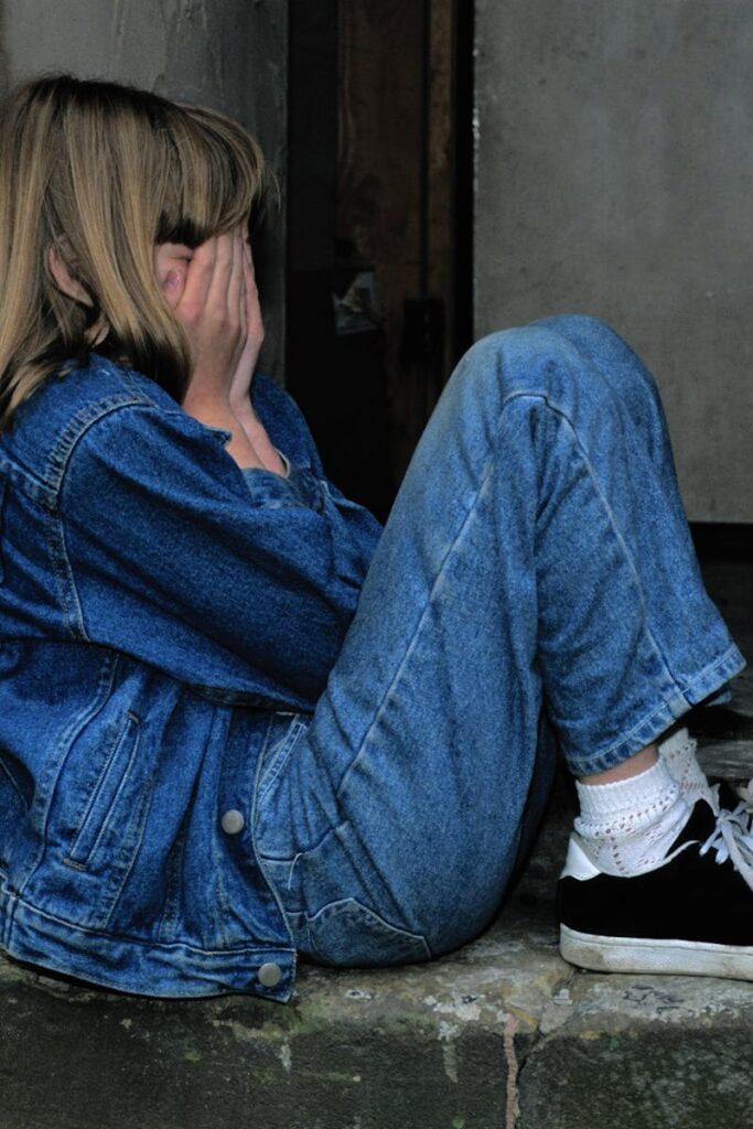 A young girl in denim sits alone in a doorway looking distressed, conveying loneliness.