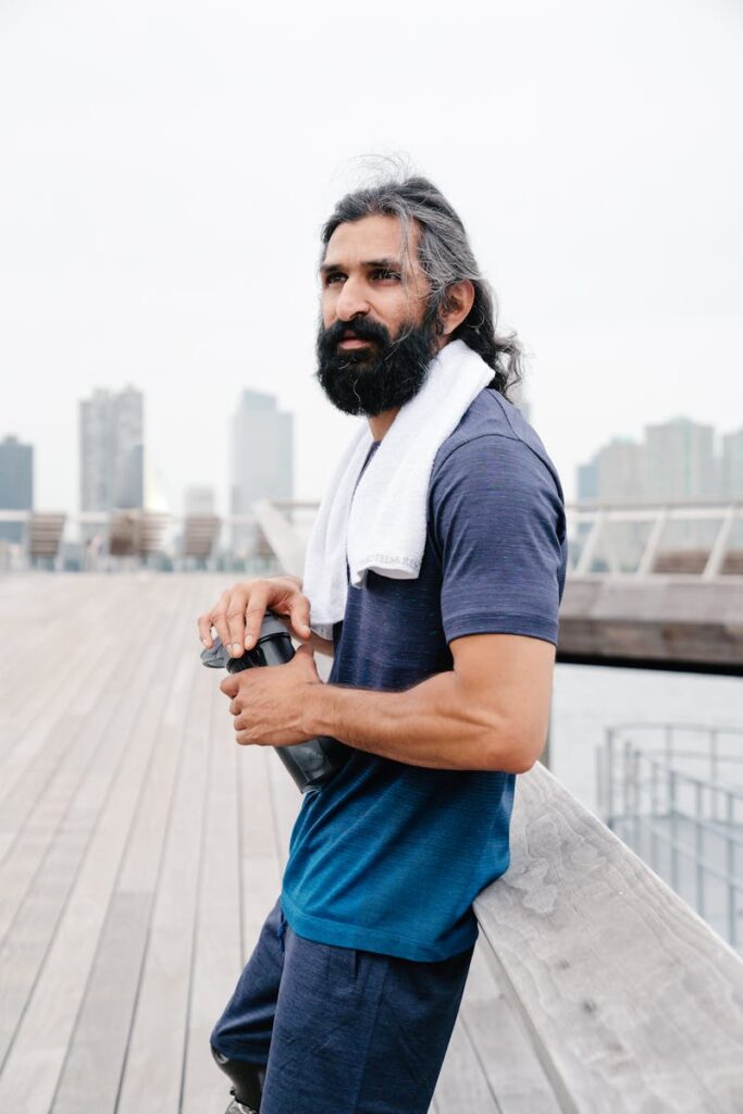 Fit man with towel and bottle relaxing after exercise by the waterfront